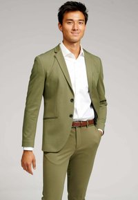 Olivengrøn dragt med en tætsiddende blazer og matchende bukser. Hvid skjorte under, brun læderbælte og knapper, glat stofstruktur.
