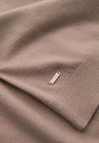 Tissu en maille marron, présentant une texture lisse et des poignets côtelés. Une petite étiquette en métal est visible près du bord inférieur.