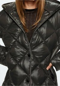 Donna che indossa un giubbotto invernale nero trapuntato con grandi cuciture a rombi e bottoni neri sopra una maglietta trasparente nera a righe.