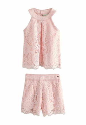 Girl with curly hair wearing a sleeveless pink lace top and matching shorts, smiling, standing with arms slightly extended.