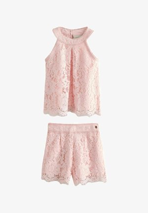 Dievča s kučeravými vlasmi oblečená v ružovom krajkovom topu bez rukávov a zodpovedajúcich šortkách, usmievajúca sa, stojaca s mierne rozpaženými rukami.