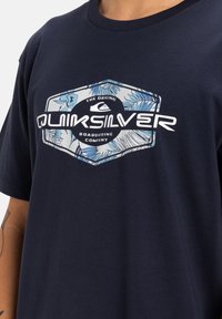 Camiseta de algodón azul marino con un gráfico hexagonal de hojas tropicales y el logo de Quiksilver en blanco y azul claro.