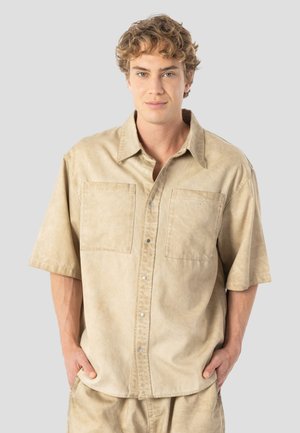 Lyhythihainen, vaaleanbeige paita, joka on valmistettu kevyestä kankaasta pehmeällä pintakuvioinnilla. Paidassa on kaksi etutaskua ja hopeiset napit napituslistassa.