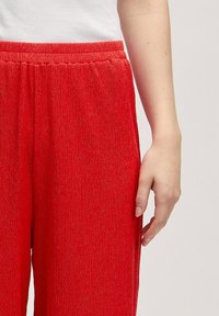 Rode, gestructureerde broek met een elastische tailleband, gekenmerkt door verticale strepen en een losse pasvorm. Hand gedeeltelijk zichtbaar voor de schaal.