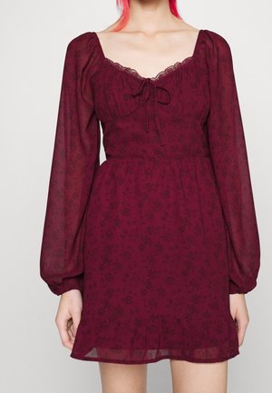 Bourgondische bloemenjurk met lange doorschijnende pofmouwen, vierkante halslijn, kanten afwerking en strikdetail op de borst, gedragen door een persoon met roze haar.