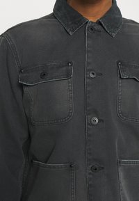Sort denimjakke med kordfløjelkrave, to brystlommer med knaplukninger og en knaplukket front. Struktureret stof med mat finish.
