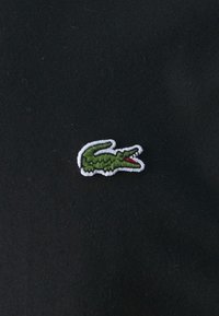Embroidered green crocodile logo on black fabric; logo features white outline, with a red accent in the mouth. Smooth texture.