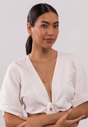 Weiße Leinenbluse mit kurzen, weiten Ärmeln, die in der Taille gebunden ist. Goldene Halskette mit einem Strahlenanhänger. Sanfte Haut, lange dunkle Haare.