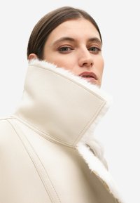 Giacca in pelle color crema con colletto alto bordato in finta pelliccia bianca, cuciture a vista e un design strutturato e angolare.