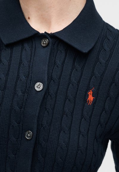 Πολό μπλούζα σε ναυτικό μπλε με πλεκτό σχέδιο καλωδίου, γιακά, τρία κουμπιά και μικρό κόκκινο κεντημένο λογότυπο αλόγου με αναβάτη στο στήθος.