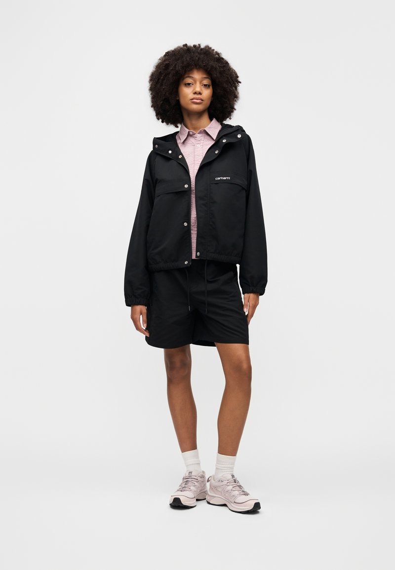 Giacca nera corta con cappuccio, dotata di tasca frontale e bottoni a pressione, indossata sopra una camicia rosa chiaro con bottoni e pantaloncini neri.