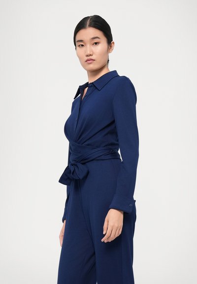 Marineblå jumpsuit med lange ærmer, krave og bindebånd i taljen. Fremstillet af blødt stof med knapdetaljer ved manchetterne.