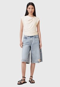 Crème top met korte mouwen en een gedraaide voorkant, gecombineerd met versleten blauwe denim Bermuda shorts en zwarte platte sandalen.