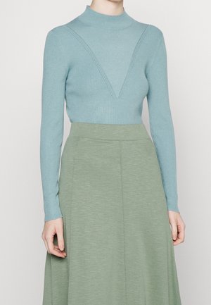 Helles blaues geripptes Sweatshirt mit hohem Kragen und V-förmigem Designdetail, kombiniert mit einem glatten, knielangen grünen Rock.