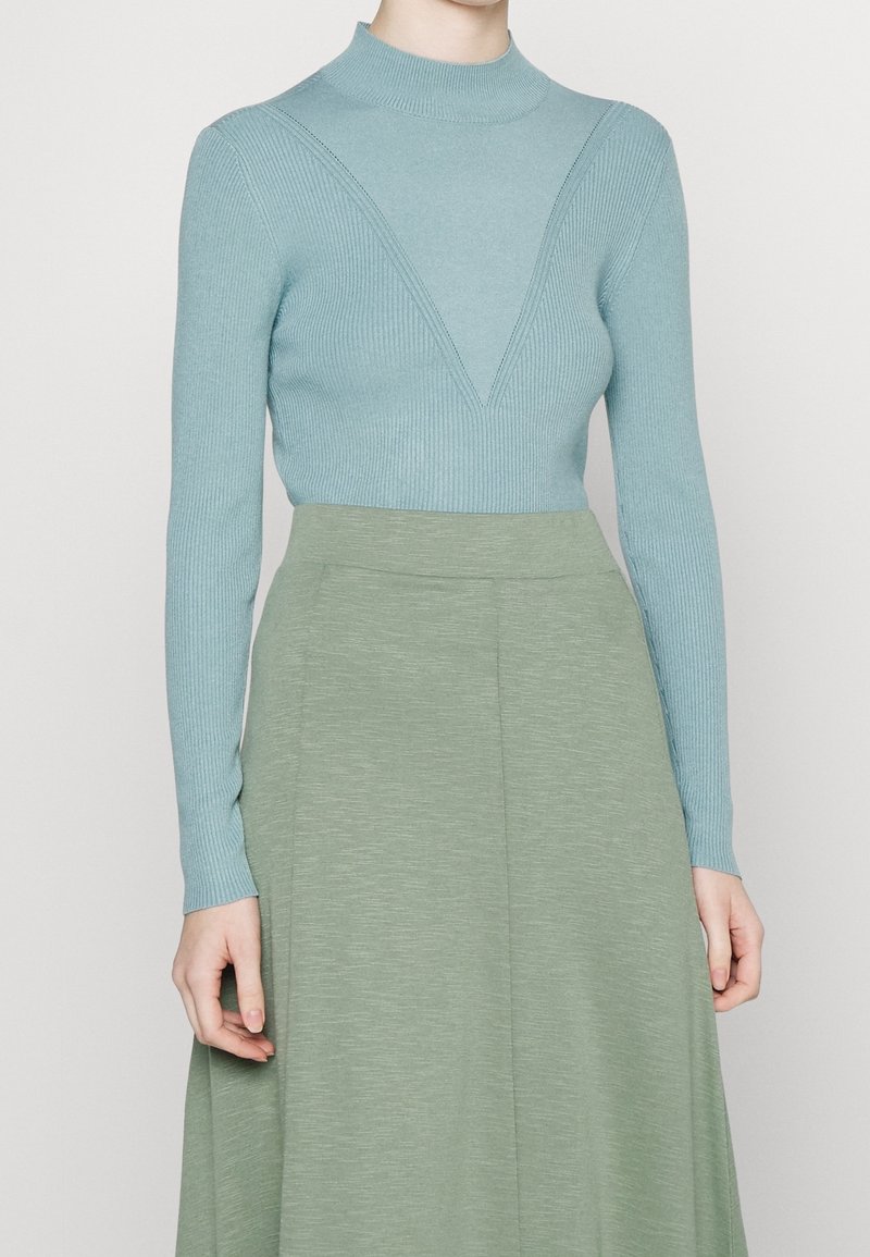 Helles blaues geripptes Sweatshirt mit hohem Kragen und V-förmigem Designdetail, kombiniert mit einem glatten, knielangen grünen Rock.