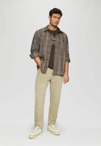Gray plaid shirt with buttoned front, worn over a dark brown t-shirt. Khaki trousers and white sneakers complete the outfit.
