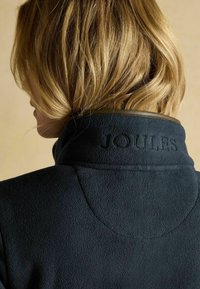 Tmavomodrá fleecová bunda s vysokým golierom a textúrovaným povrchom. Na zadnej strane sa nachádza vyrazený názov značky "Joules" v decentnom dizajne.