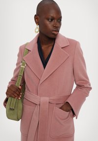 Rosa ullmixkappa med midjebälte, spetsiga slag och två fickor; kombinerad med en grön mockaaxelväska och guldörhängen i hoop-stil.