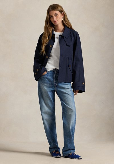 Μπουφάν από καμβά σε ναυτικό μπλε με μεγάλες μπροστινές τσέπες, κλείσιμο με κουμπιά και φαρδιά μανίκια, συνδυασμένο με ανοιχτόχρωμο τζιν και μπλε παπούτσια τύπου slip-on.