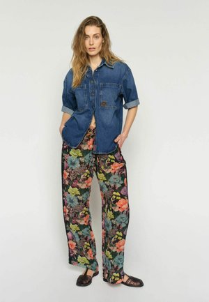 Femeie purtând o cămașă din denim rulată, pantaloni largi cu imprimeu floral în mai multe culori și pantofi negri planați, împletiți, stând în picioare lângă un perete simplu.