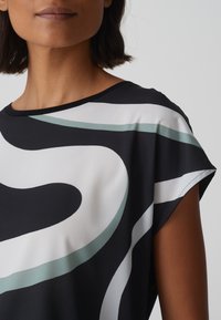 Zwart-wit top met abstracte draaikolken in licht teal. De stof heeft een gladde textuur, een ronde halslijn en korte mouwen.