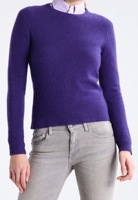 Persona che indossa un maglione lungo viola sopra una camicia colletto viola chiaro, abbinato a jeans grigi chiari, in piedi con le braccia rilassate.