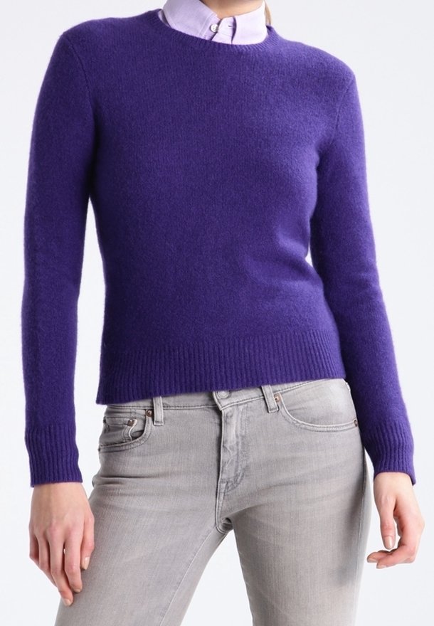 Persona che indossa un maglione lungo viola sopra una camicia colletto viola chiaro, abbinato a jeans grigi chiari, in piedi con le braccia rilassate.