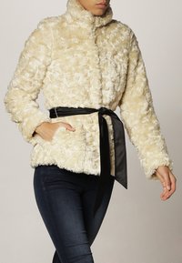 Creme faux fur jakke med høj krave, tekstureret overflade og sort bælte i taljen. Har to sidelommer og lange ærmer.