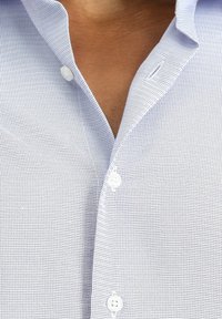 Camicia azzurra chiara a maniche corte con un piccolo motivo a quadri, dotata di un collo a punta e bottoni bianchi per la chiusura. La texture appare liscia.