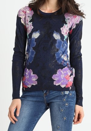 Woman wearing navy blue long-sleeve sweater with large pink and purple floral print paired with blue jeans featuring embroidered flowers on the thigh.