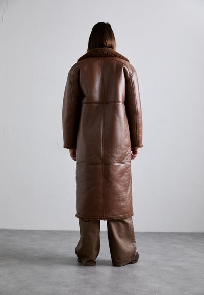 Persona vista da dietro che indossa un lungo cappotto di pelle marrone con il colletto e i polsini di pelliccia, in piedi su un pavimento grigio di fronte a un muro semplice.