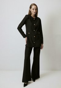 Black tailored blazer with gold buttons, featuring a round neckline and long sleeves, paired with black flared trousers and pointed black shoes.