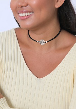 Black choker with a silver geometric pendant and two silver accents. Model wears a light yellow, ribbed top with a v-neckline.