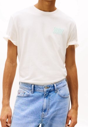 Homme portant un t-shirt blanc à manches retroussées avec un logo brodé bleu clair et un jean en denim bleu clair taille haute.