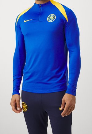 Mann trägt blaues langärmeliges Sportoberteil mit gelben Akzenten und schwarze Hose, beide mit Nike-Logo und gelbem kreisförmigem Emblem.
