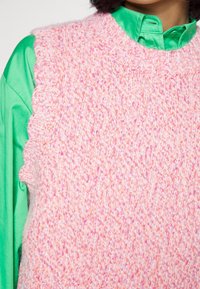 Lähellä näkyvä pinkki ja oranssi neulottu ilman hihoja oleva villapaita, joka on puettava kirkkaan vihreän satiinipainon päällä.