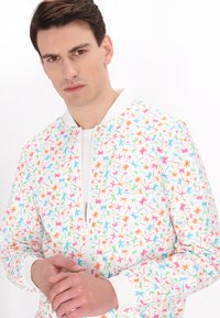 Witte bomberjack met een kleurrijk patroon van bladeren en bloemen in roze, blauw en groen. Voorzien van een ritssluiting aan de voorkant en geribde manchetten.