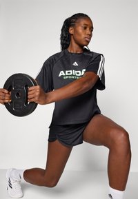 Juoda sportinė marškinė su žalia "ADIDAS" logotipu, baltomis juostelėmis ir juodais šortais. Modelis laiko apvalų juodą svorį, esančiame puolimo pozicijoje.