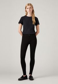 Zwarte fitted jeans met een hoge taille, gecombineerd met een zwart T-shirt met korte mouwen. Het model draagt zwarte instappers. Gladde stoftextuur.
