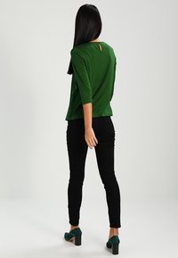 Una persona con lunghi capelli scuri è di spalle, indossa una maglietta verde con un taglio sulla schiena e pantaloni neri, abbinati a tacchi verdi.