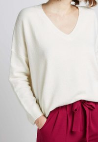Pull blanc à col en V en laine douce, avec des manches amples et une coupe décontractée, associé à un pantalon rose taille haute avec une taille nouée.