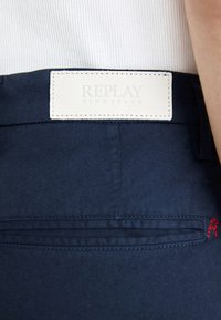 Navy blue jeans with a white leather waistband patch embossed with "REPLAY BLUE JEANS." Features a back pocket with red stitching detail.