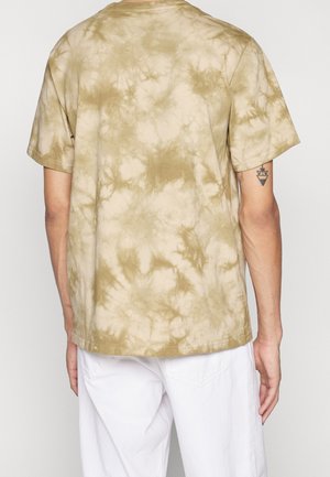 Person, der ein beige- und hellbraunes Batik-T-Shirt mit kurzen Ärmeln und weiße Hose trägt, von hinten gezeigt, mit sichtbarem Arm-Tattoo am rechten Arm.