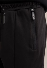 Pantaloni sportivi neri con tasca con zip e vita regolabile con cordino. Il materiale è liscio con una finitura leggermente testurizzata.