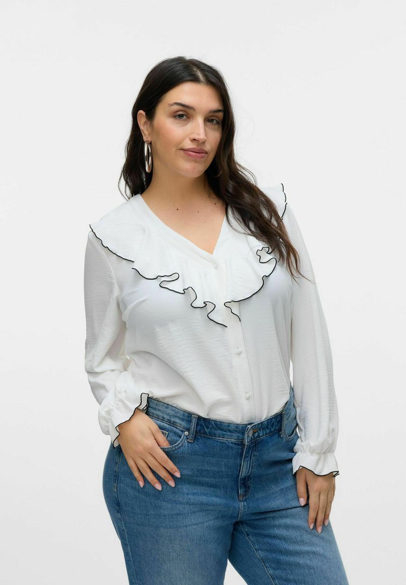 Blouse blanche à boutons avec des accents de volants noirs le long du col et des manches, associée à un jean en denim bleu foncé. Texture lisse.