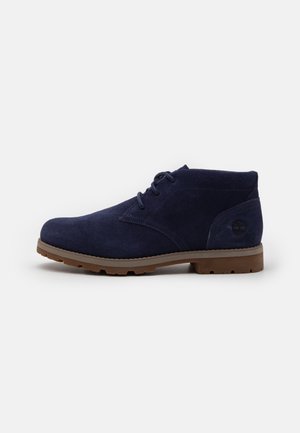 Granatowy zamszowy botek za kostkę z brązową gumową podeszwą i sznurowadłami, widok z boku, pokazujący wytłoczone logo Timberland w pobliżu pięty.