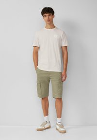 T-shirt en coton beige clair avec un petit logo, associé à un short cargo vert olive avec des poches latérales, et des baskets blanches avec des accents beiges.