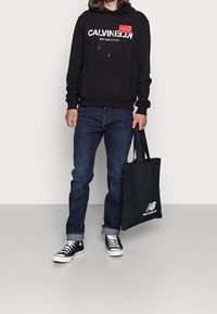 Sudadera negra con logo blanco de "CALVIN KLEIN", vaqueros, zapatillas negras y una bolsa de mano negra con el logo blanco de "new balance".