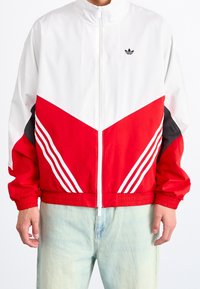 Red and white track jacket with a high collar, zip closure, chevron design, and three white stripes on each sleeve. Black accents on sides.