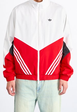Veste de piste rouge et blanche avec un col haut, fermeture éclair, design en chevrons et trois bandes blanches sur chaque manche. Accents noirs sur les côtés.